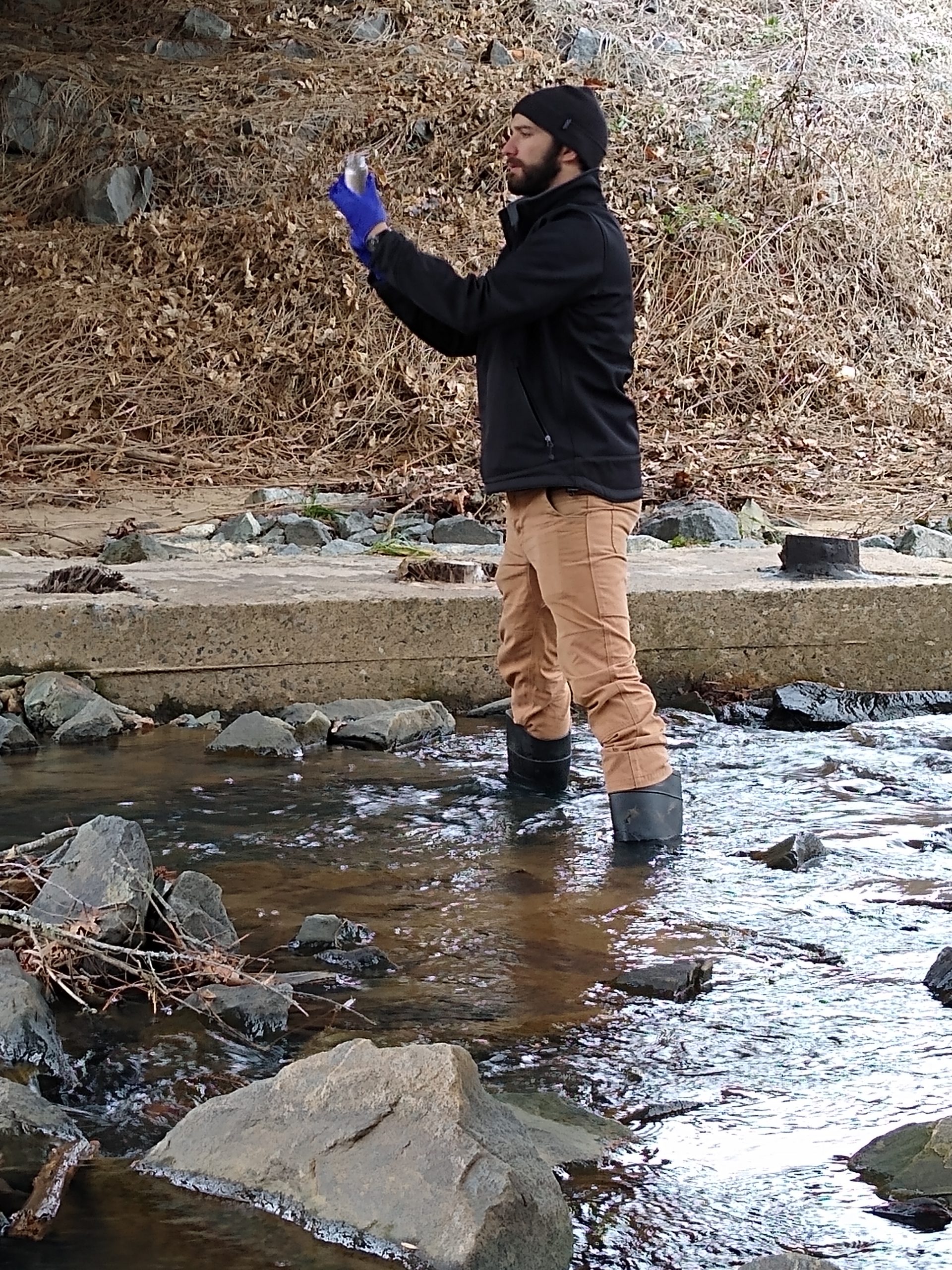 A man is standing in a river, with knee high boots on to protect from the water, holding a clear jar to collect a sample of water.