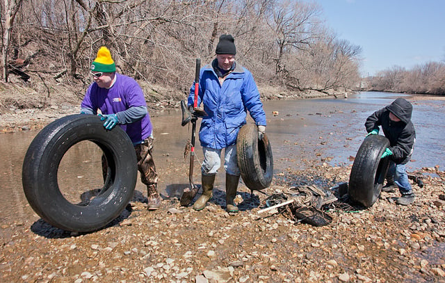 Three people roll large tires away from a riverbank.